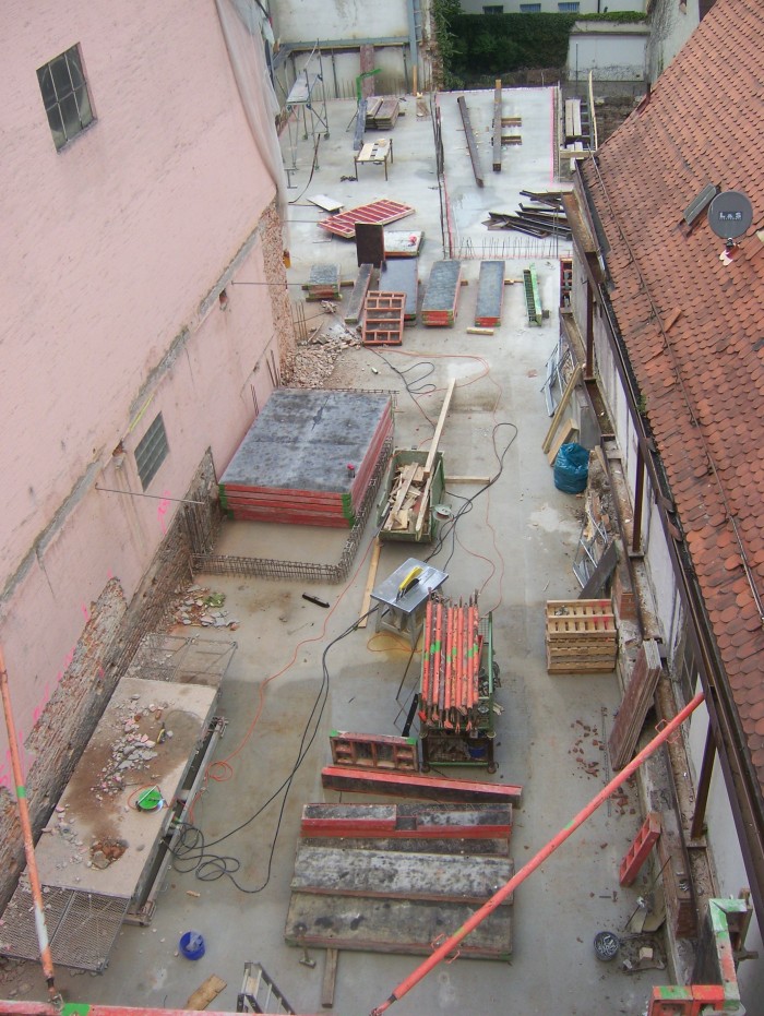 Blick von oben auf das im Bau befindliche 1. Obergeschoss des Neubaus, auf dem Boden stehen bzw. liegen verschiedene Baumaterialien.