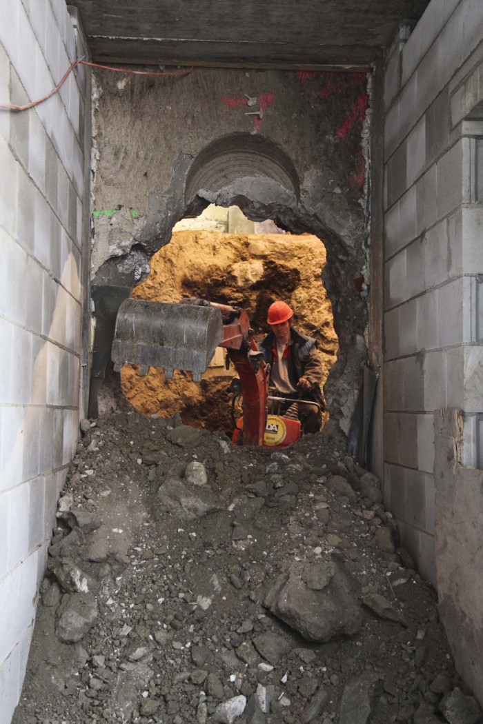 Ein großes Loch in der Mauer, davor ein Geröllhaufen, dahinter ein Bagger, welcher von einem Bauarbeiter mit Helm bedient wird.