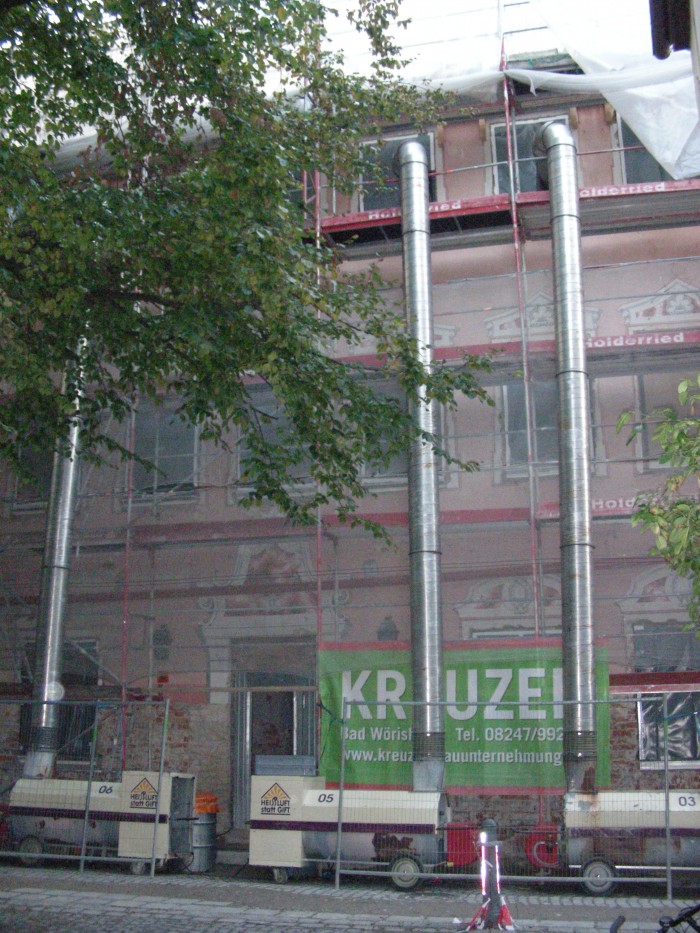 Blick auf die Fassade des Stadtmuseums, welche eingerüstet und mit einem Netz davor versehen ist, in drei der oberen Fenster führen silberne Rohre, welche unten jeweils mit einer Maschine verbunden sind, an der Fassade unten rechts befindet sich ein Werbeplakat der Firma Kreuzer