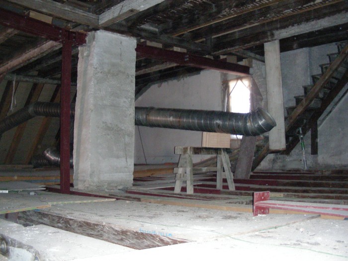 Blick in den Dachstuhl der Altbaus, von der linken Seite führt ein silbernes Rohr in die Mitte des Raumes, im Hintergrund ein Fenster und eine Treppe nach oben.