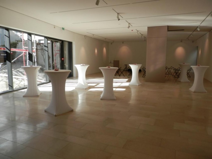 Blick in den Sonderausstellungsraum mit sieben Stehtischen ausgestattet, links Blick durchs Fenster in den verschneiten Museumshof, die Sonne scheint in den Ausstellungsraum, im Hintergrund Bestuhlung