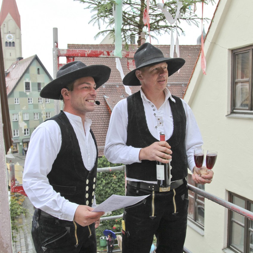 zwei Zimmerer auf einem Gerüst, einer verliest den Richtspruch, der andere mit einer Flasche und zwei Gläser Sekt, im Hintergrund der Turm der Martinskirche