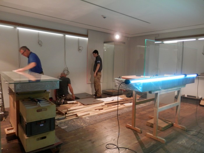 Im Hintergrund bauen drei Männer Vitrinen ein, auf der linken Seite eine Werkbank auf der eine Glasplatte liegt, im Vordergrund eine UV-Lampe