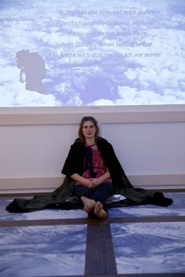 Eine Frau (Lisa) im Umhang sitzt auf dem Boden und schaut in die Kamera. Hinter ihr die vom Beamer an die Wand projezierte Präsentation von Sophie von La Roche.
