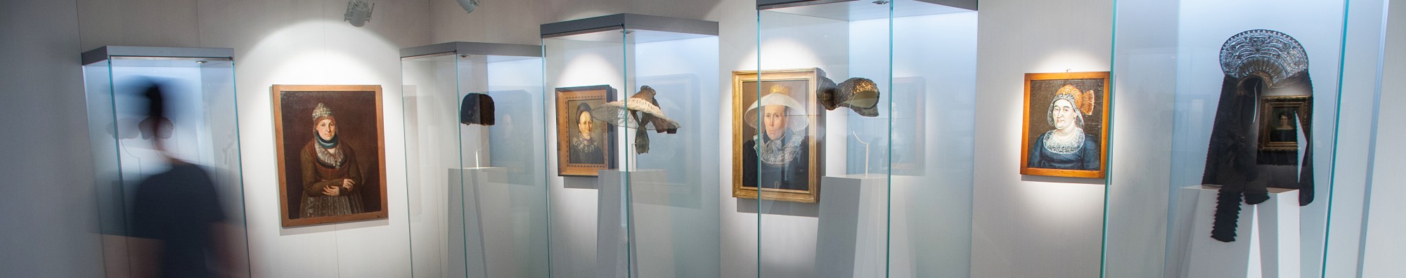 Blick in den Haubenraum: vier Vitrinen, in denen verschiedene Hauben ausgestellt sind. Dazwischen Gemälde von Frauen, die mit diesen/ähnlichen Hauben zu sehen sind.