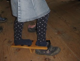 Mädchen zieht sich mit einem &quot;Stiefelknecht&quot; den Schuh aus. Ein Stiefelknecht ist ein Holzbrett mit hufeisenförmigem Ausschnitt, in den man seinen Fuß steckt und so den Schuh abstreifen kann.