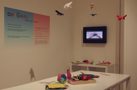 Blick in die Ausstellung: Auf einem Tisch liegen bunte Origami, in der Luft hängen ebenfalls Origami. Im Hintergrund ist ein Bildschirm, der eine Bastelanleitung zeigt.
