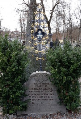 Grab des Pfarrers Richard Wiebel: Grabstein mit Eisenkreuz darauf, links und rechts zwei kleine Büsche. Dahinter ein der Friedhof und ein paar Bäume