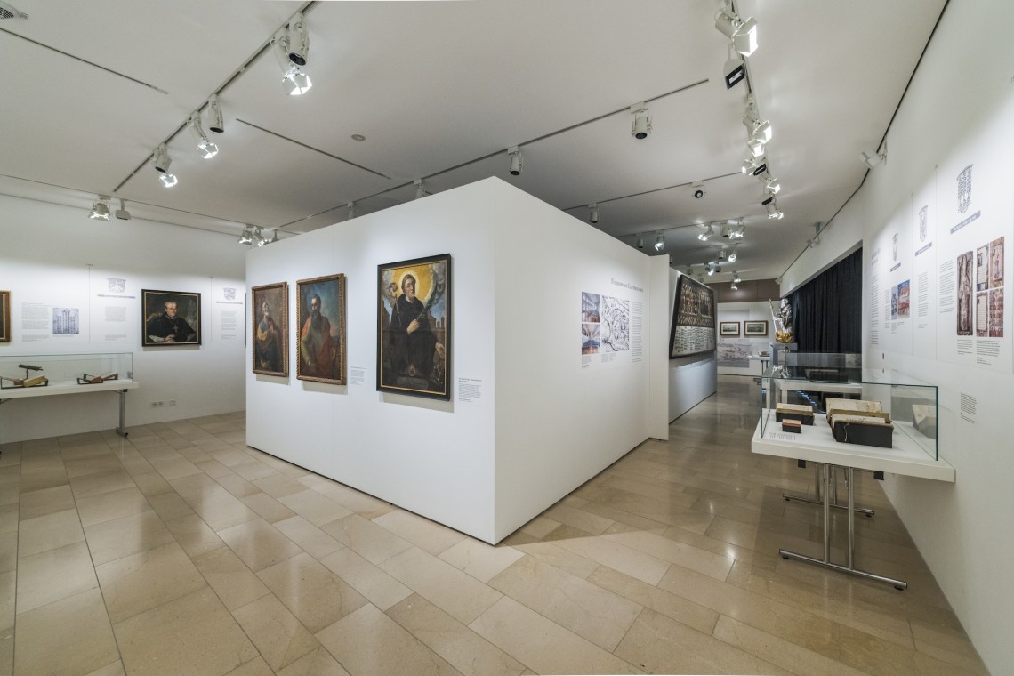 Blick in die Ausstellung: Gemälde an den Wänden, Glasvitrinen