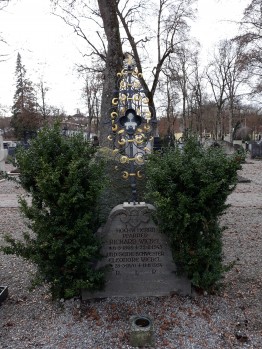 Grab des Pfarrers Richard Wiebel: Grabstein mit Eisenkreuz darauf, links und rechts zwei kleine Büsche. Dahinter der Friedhof und ein paar Bäume