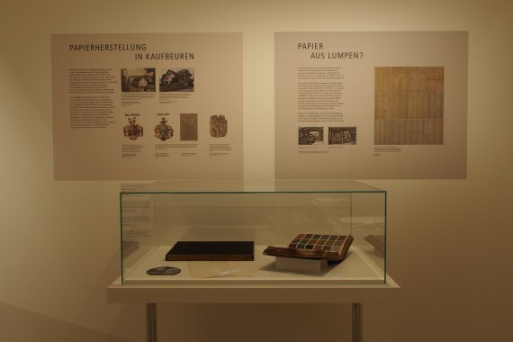 Blick in die Ausstellung: eine Glasvitrine mit Inhalt, dahinter 2 Informationstafeln über die Papierherstellung und &quot;Papier aus Lumpen?&quot;