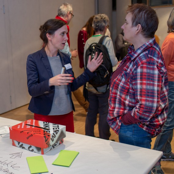 Museumsleiterin Petra Weber im Gespräch mit einer Frau, im Hintergrund mehrere Menschen, die sich ebenfalls unterhalten. Im Vordergrund eine Feedback-Box auf einem Tisch