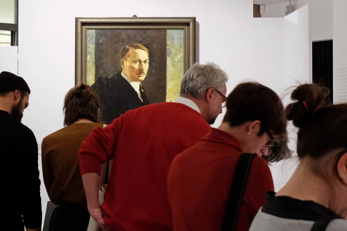 Hintergrund: Porträt von einem Mann, der Hitler vom Aussehen sehr ähnelt (Scheitelfrisur, typischer Schnauzbart). Davor Menschen, die sich die Ausstellung ansehen.