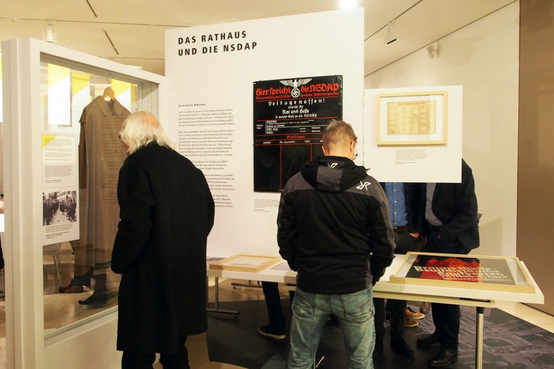 Zwei Besucher sehen sich die Themensinsel &quot;Das Rathaus und die NSDAP&quot; an. Links hängt ein brauner BDM-Mantel, geradeaus ist ein Infotext und ein Plakat abgebildet.