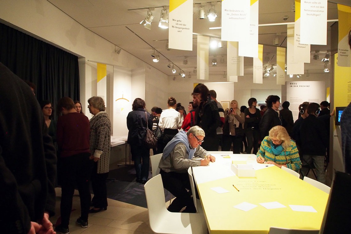 Der Ausstellungsraum ist voller Menschen. Zwei Besucher:innen schreiben eine Frage an einem Tisch auf. 