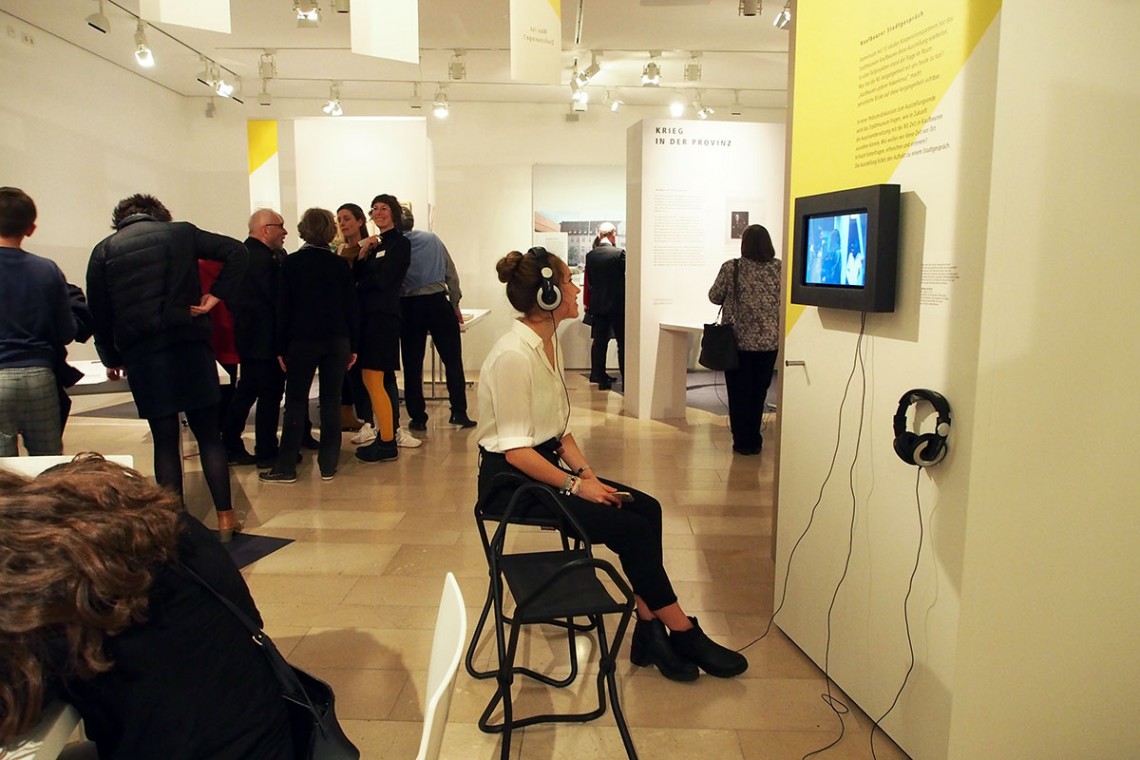 Eine Besucherin schaut sich mit Kopfhörern einen Film auf einem kleinen Bildschirm an. Im Hintergrund begutachten weitere Besucher verschiedene Themeninseln.