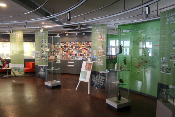 Blick in den Infopoint Museen und Schlösser in Bayern. Rechts und links Banner der &quot;Familienmuseen Allgäu&quot;, in der Mitte ein Regal gefüllt mit Flyer