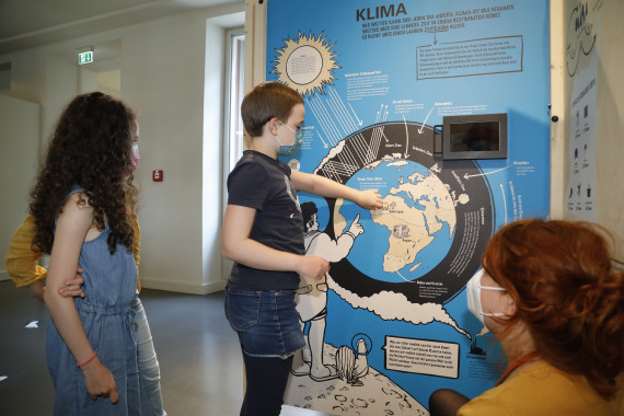 einer Station mit der Aufschrift Klima, links daneben zwei Schülerinnen, rechts eine Frau 