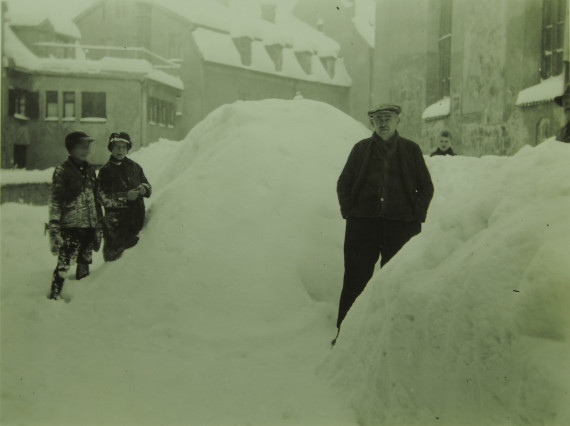 im Hintergrund Häuser, im Vordergrund mannshohe Schneehaufen, links stehend zwei Buben und links ein Mann, schwarz/weiß-Foto aus dem Jahr 1931
