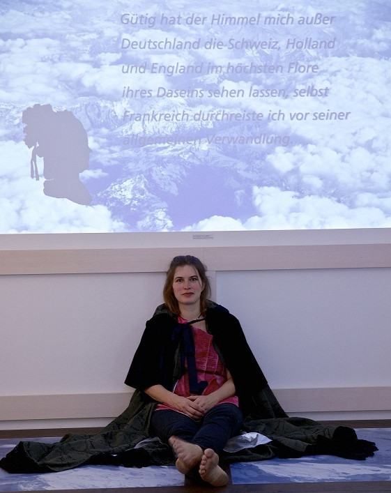 Eine Frau (Lisa) im Umhang sitzt auf dem Boden und schaut in die Kamera. Hinter ihr die vom Beamer an die Wand projezierte Präsentation von Sophie von La Roche.