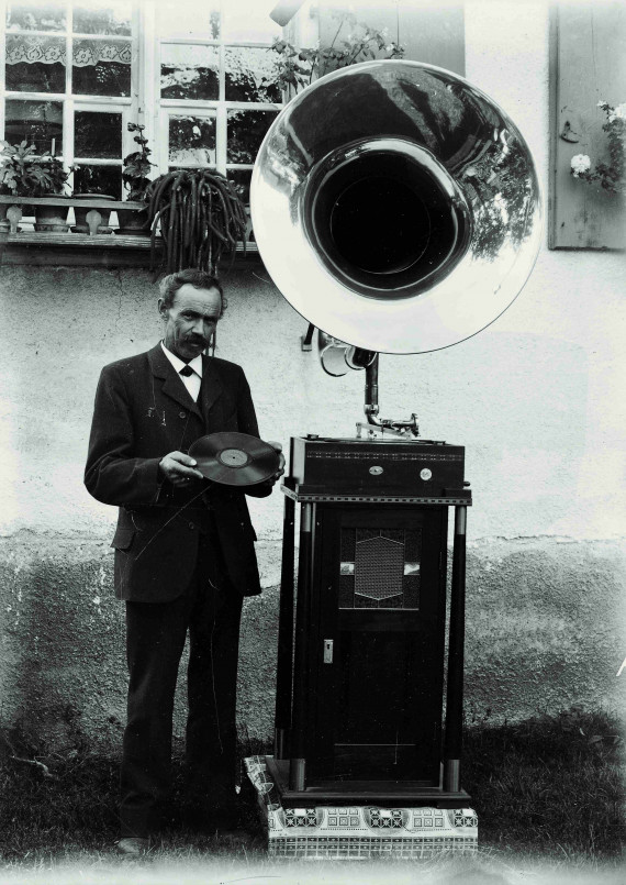 Mann steht neben einem großen Grammophon vor dem Haus und hält eine Schallplatte in der Hand.