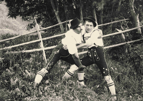 Zwei Männer in Lederhosen posieren draußen auf einer Wiese, als würden sie miteinander rangeln.