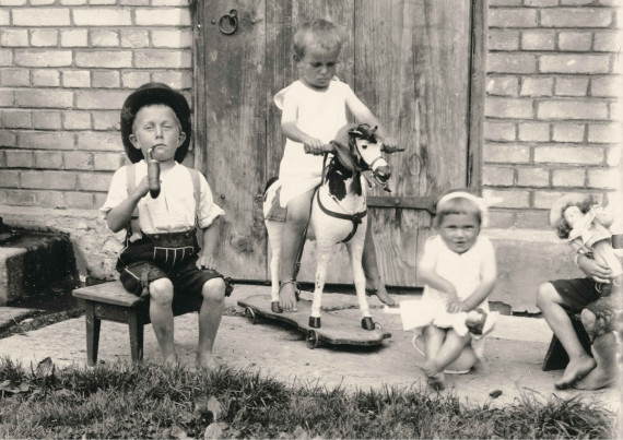 Drei spielende Kinder vor einer Holztüre, links ein Bub mit einer Pfeife, in der Mitte ein Kind auf einem Schaukelpferd.